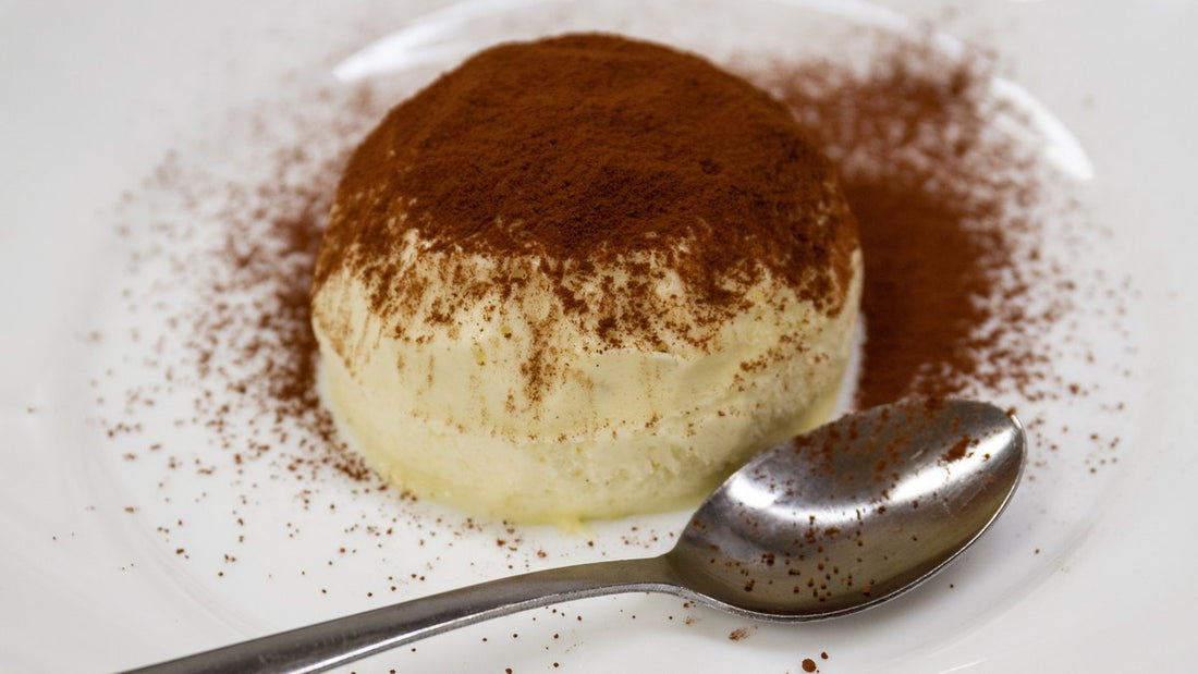 Tiramisu senza uova IN-VECE spolverato di cacao su piatto bianco, con cucchiaino