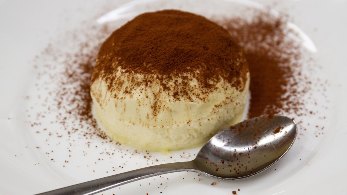 Tiramisu senza uova IN-VECE spolverato di cacao su piatto bianco, con cucchiaino